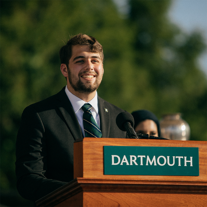 Jared speaking at a Dartmouth podium with trees in the background.